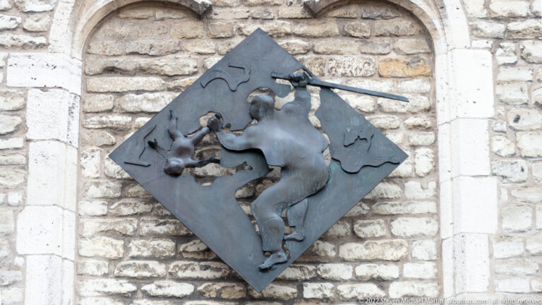 Dark metal relief of a horse and rider mounted on a diamond-shaped plate against pale stone masonry.