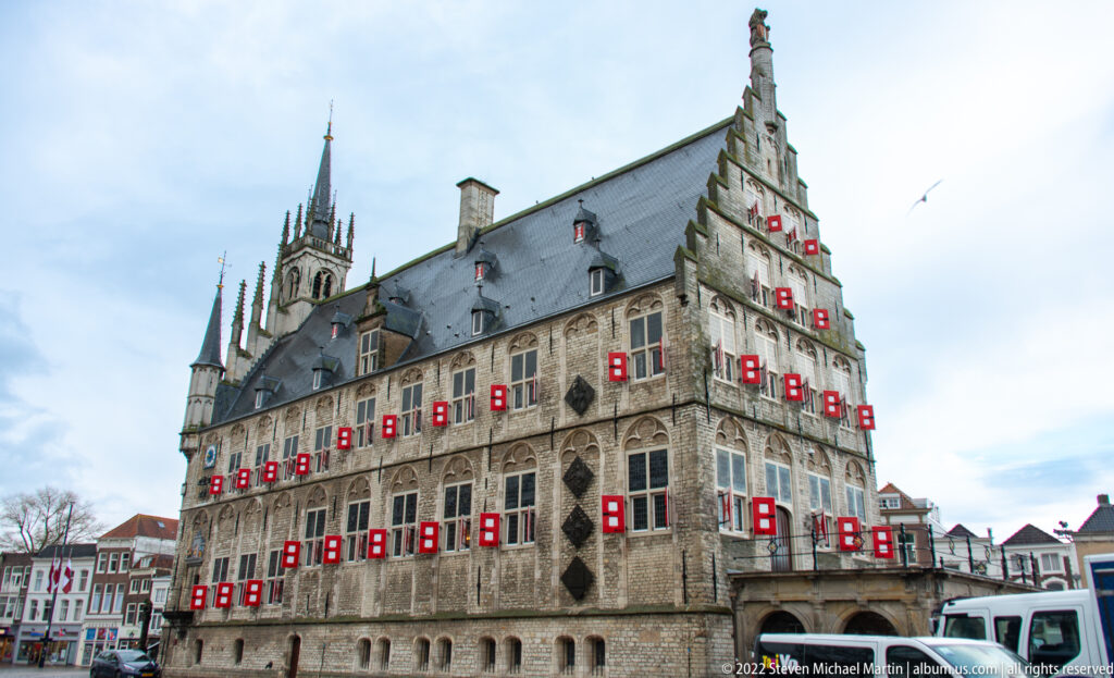Oblique view of Gouda Town Hall showing stepped gable, arcaded ground floor, steep roof, pinnacles, and red-white shutters.