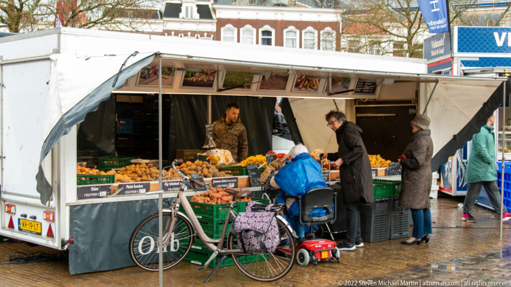 Outdoor produce stall in Gouda with vendor, shoppers, crates of fruit and vegetables, and a bicycle beside the counter.