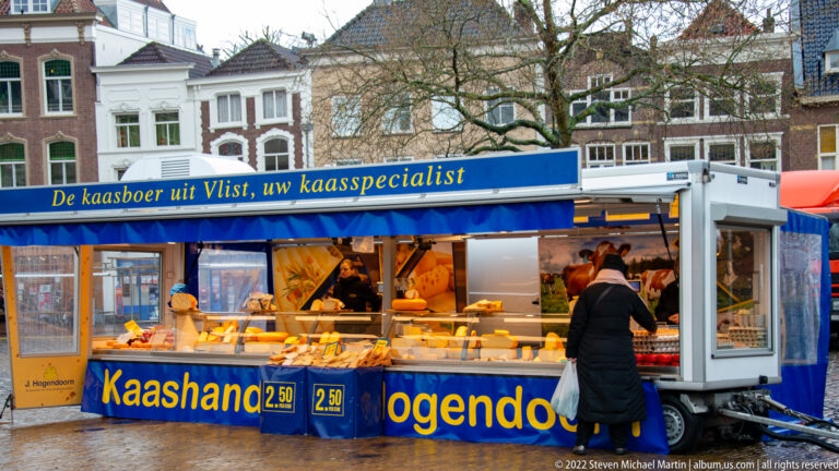 Blue-and-white market stall selling cheese, with Dutch signage, refrigerated display, vendor area, and a customer at the counter.