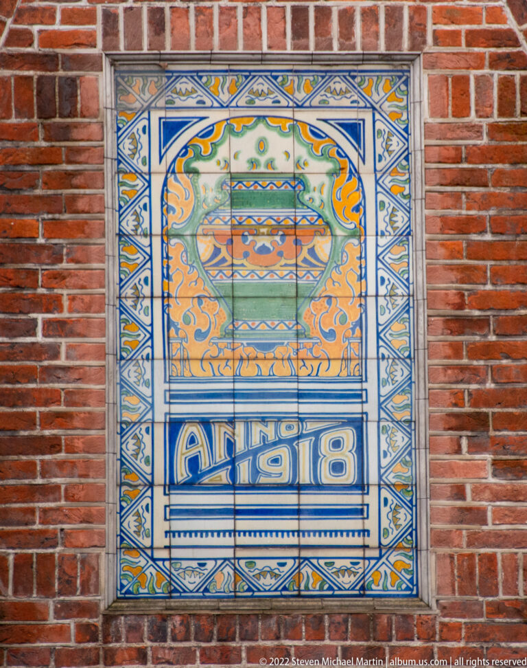 Decorative glazed tile panel in a brick wall with a vessel motif, ornate border, and the date “Anno 1918.”