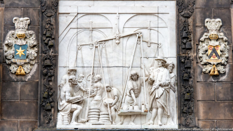 White stone relief on the Gouda Waag showing figures around a large balance, flanked by heraldic shields.
