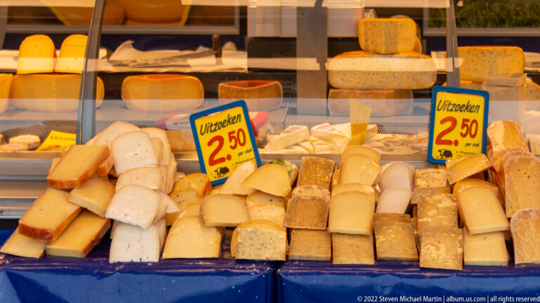 Market display piled with assorted cheese wedges and small wheels beside handwritten Dutch price signs reading “Uitzoeken 2,50.”