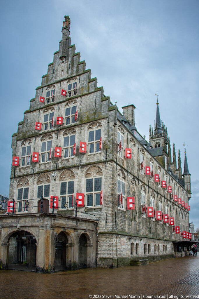 Side view of Gouda Town Hall with stepped gable, ground-floor arches, red-white shutters, steep roof, and pinnacled tower.