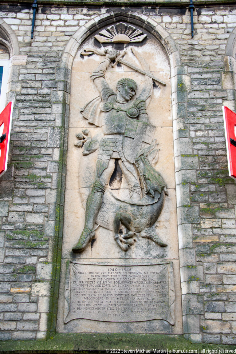 Weathered stone relief of Saint George on horseback spearing a dragon, with an inscribed plaque below.