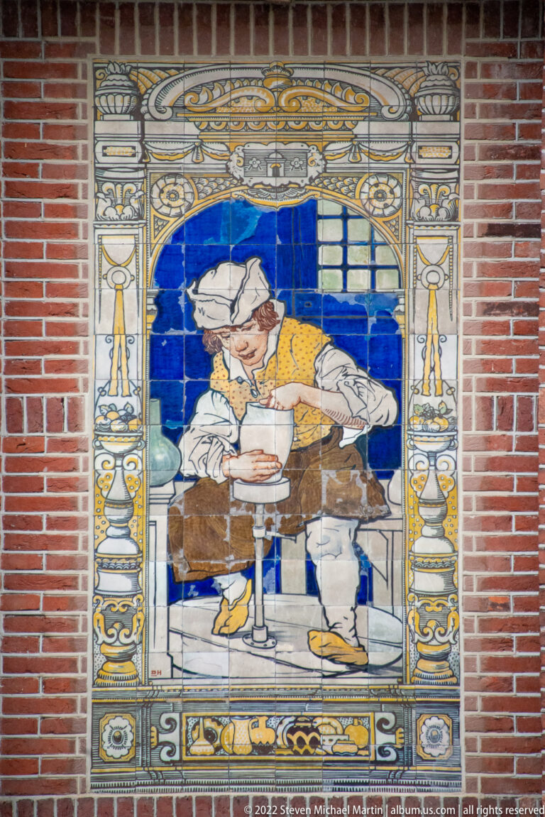 Glazed tile mural in a brick wall showing a seated male worker within an ornate architectural border.