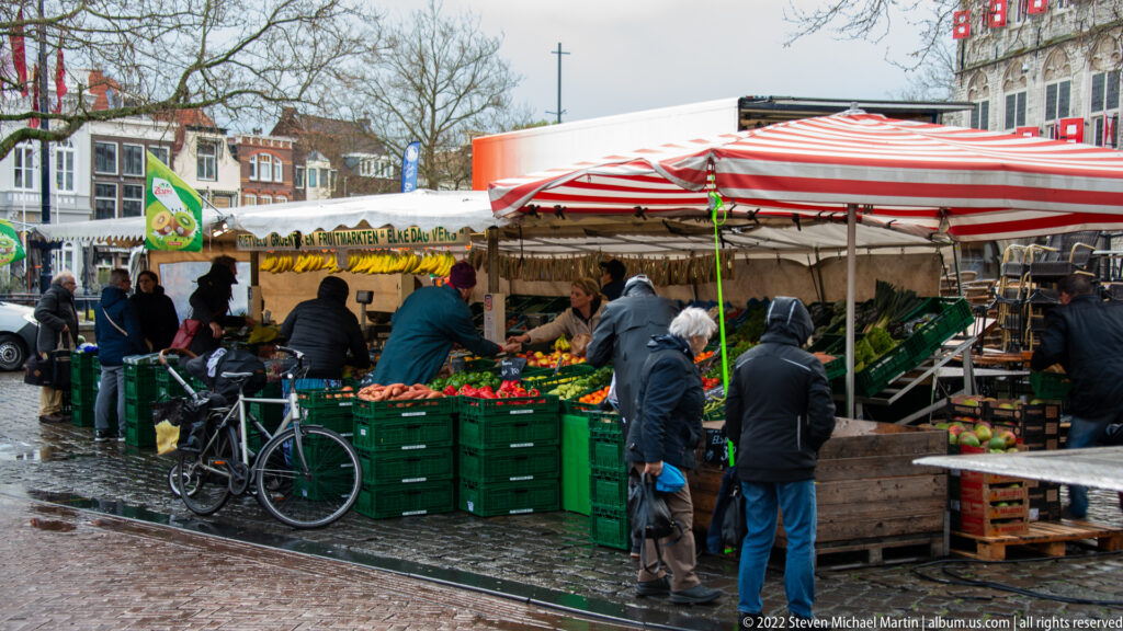 Shoppers at a produce stall under red-and-white striped canopies, with green crates, vegetables, bicycles, and wet paving.