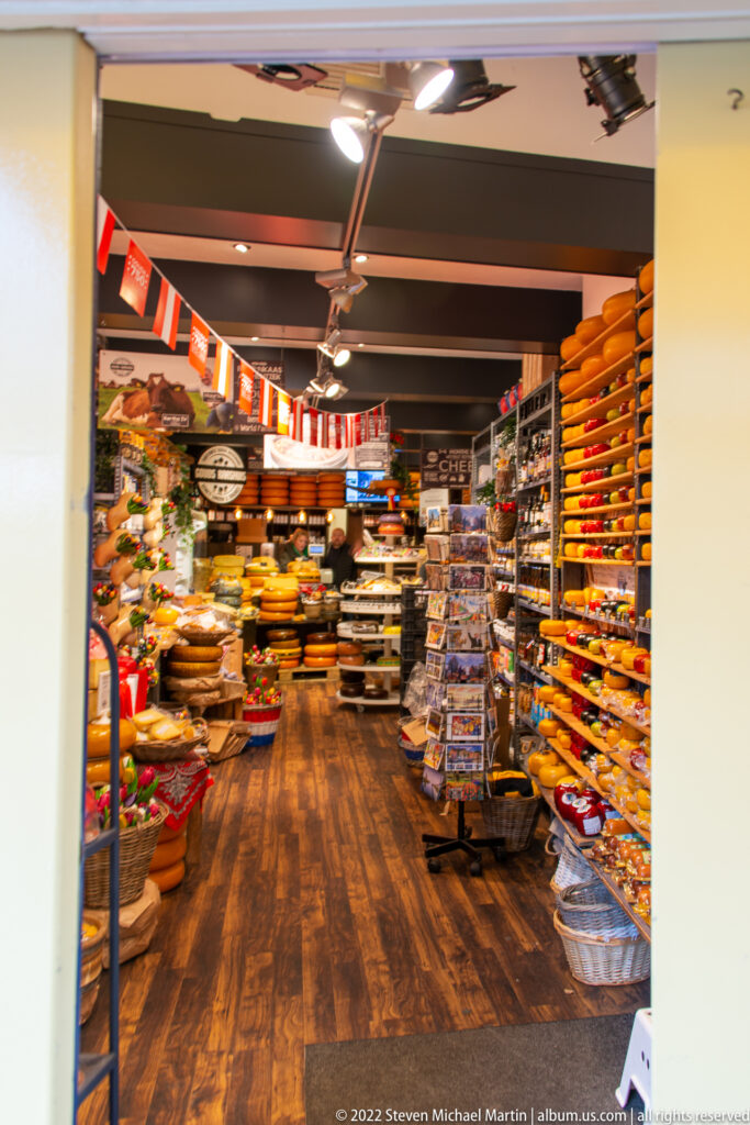 Interior of a Gouda cheese shop with yellow cheese wheels stacked on shelves and merchandise displays filling the aisle.