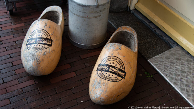 Two large weathered wooden clogs marked Gouds Kaashuis rest on brick paving outside a shop entrance.