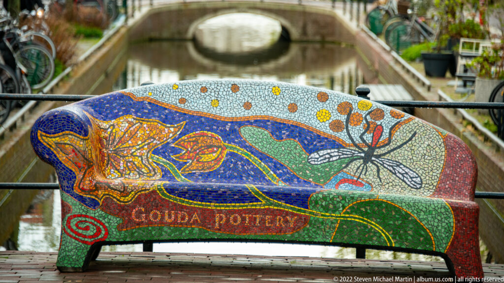 Colorful mosaic bench beside a canal, with the words Gouda Pottery visible across its front.