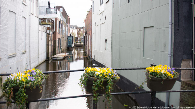 Narrow canal between close-set buildings, viewed past a railing with yellow and white flower baskets.