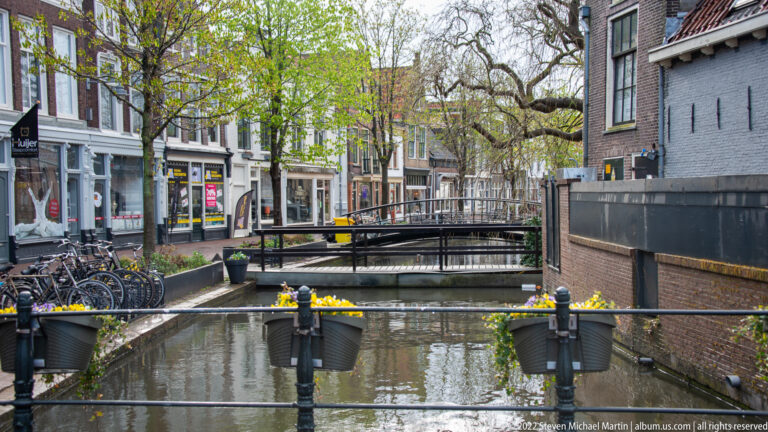Small bridge over a Gouda canal with bicycles, trees, shops, and hanging flower pots.