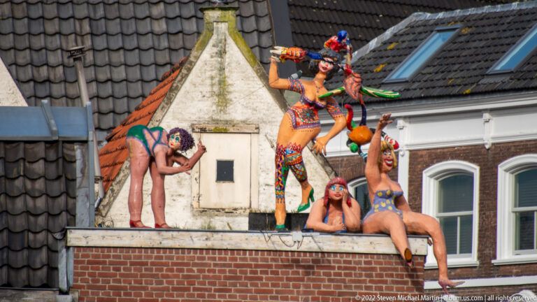 Four brightly painted human figures posed on a low rooftop among tiled roofs and brick buildings.