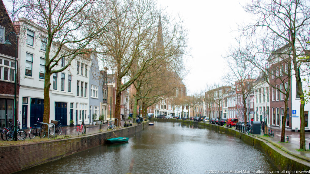 Long canal view in Gouda with brick quays, narrow houses, bicycles, leafless trees, parked cars, and a small boat.