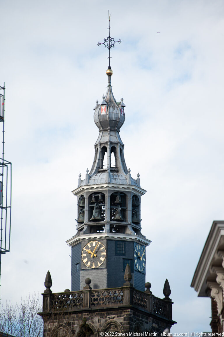 Historic tower with black-and-gold clock face, open belfry, lantern, and weather vane.