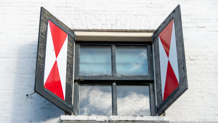 Small window in white brick wall with black-framed shutters painted in red-and-white diagonal patterns.