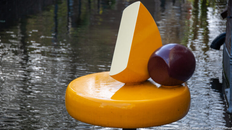 Yellow cheese-themed sculpture beside water, with a wedge and dark sphere resting on a circular base.