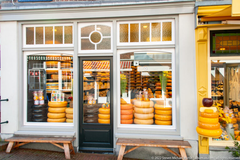 Cheese shop façade with stacked wheels visible through large windows on both sides of a dark door.