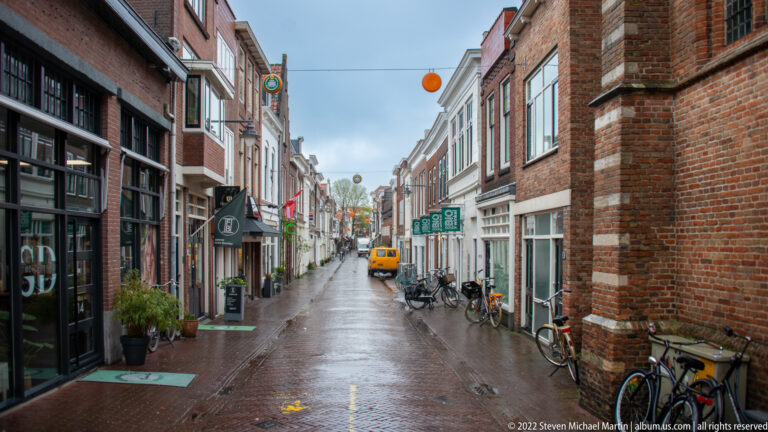 Rain-wet narrow street in Gouda with brick buildings, shops, bicycles, signs, and a small delivery vehicle.
