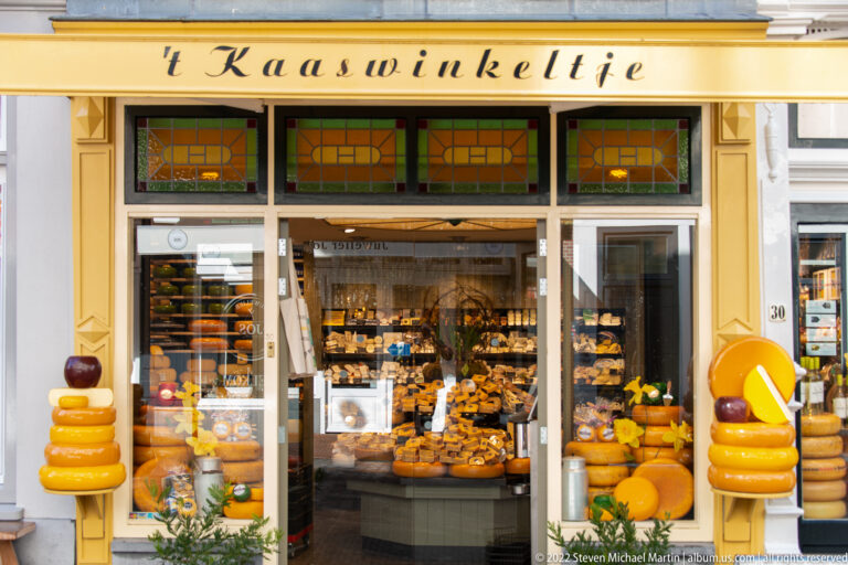 Yellow-trimmed 't Kaaswinkeltje storefront filled with cheeses and flanked by stacked cheese sculptures.