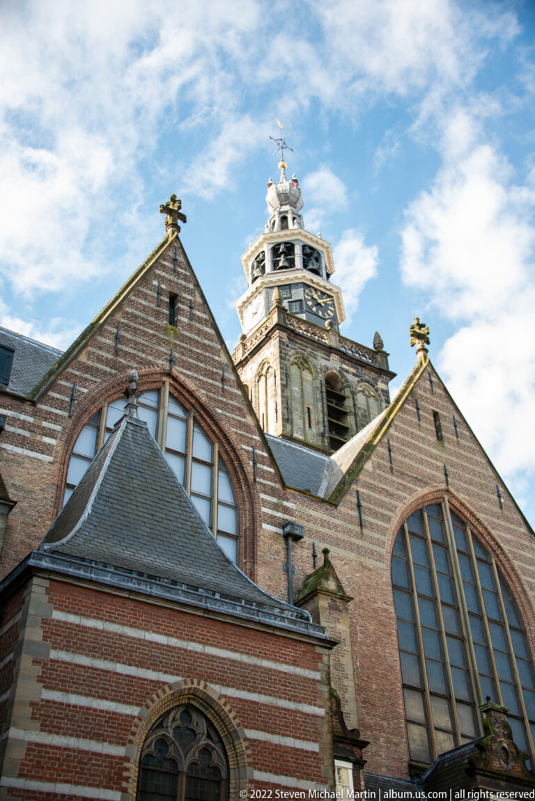Low-angle view of Sint-Janskerk brick gables, tall arched windows, and tower against a blue sky.