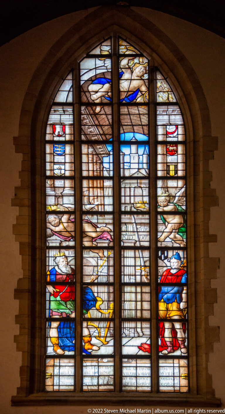 Large arched stained-glass window with architectural imagery and bands of red, blue, yellow, and green glass.
