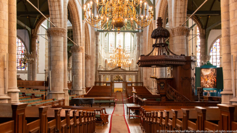 Interior of Sint-Janskerk with stone arcades, wooden pews, brass chandeliers, pulpit, and stained-glass windows.
