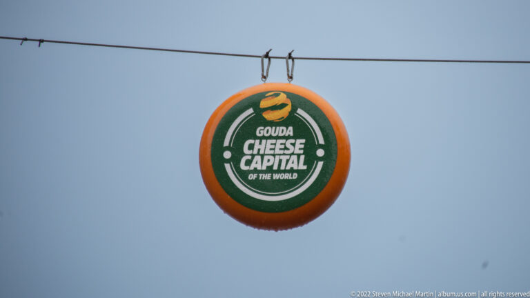 Round orange-and-green hanging sign reading “Gouda Cheese Capital of the World” against a gray sky.