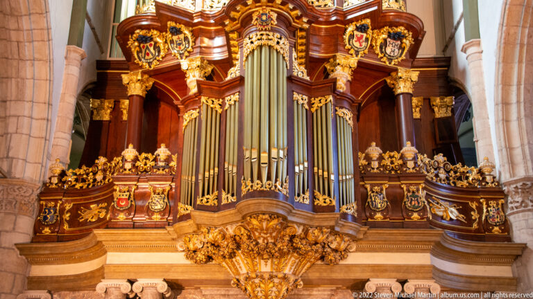 Close frontal view of an ornate church organ with gilded carving, heraldic shields, and tall silver pipes.