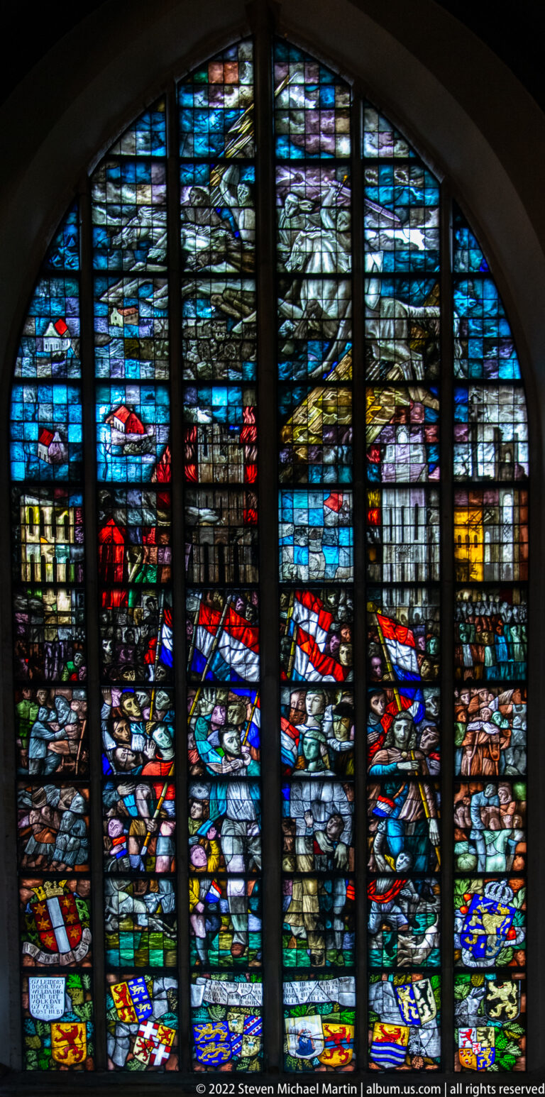 Large stained-glass window with blue narrative scenes and rows of colorful heraldic shields in Sint-Janskerk.