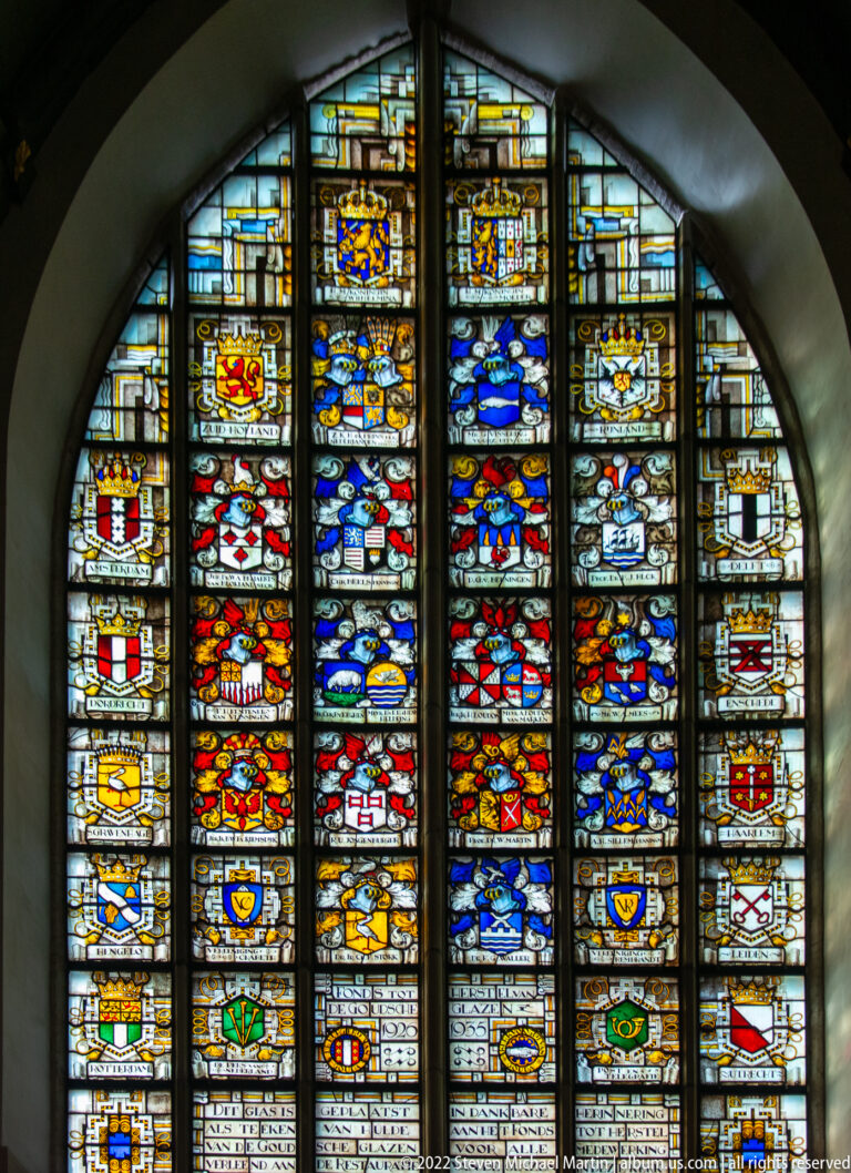 Arched stained-glass window filled with vertical rows of colorful coats of arms and small upper scenes.