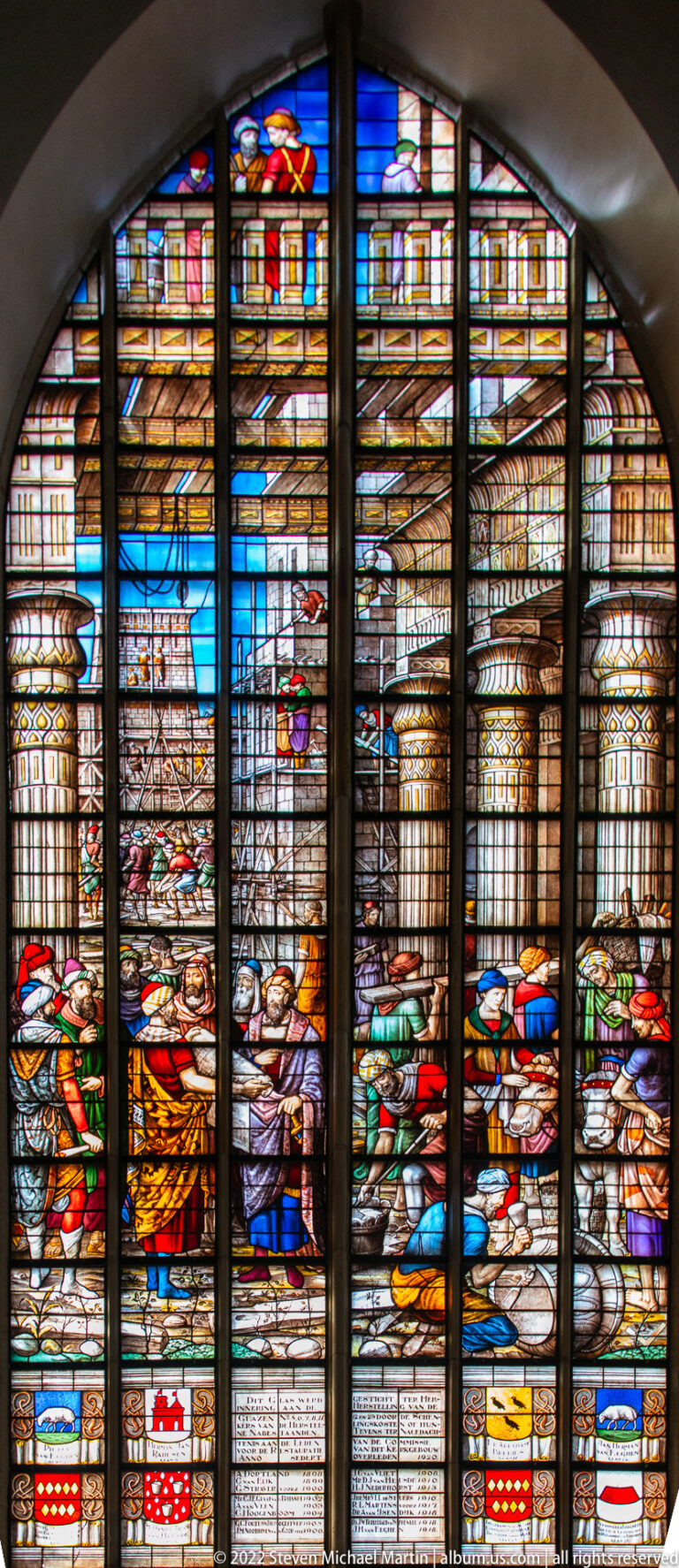 Stained-glass window with architectural scenes, standing figures, and heraldic shields below.