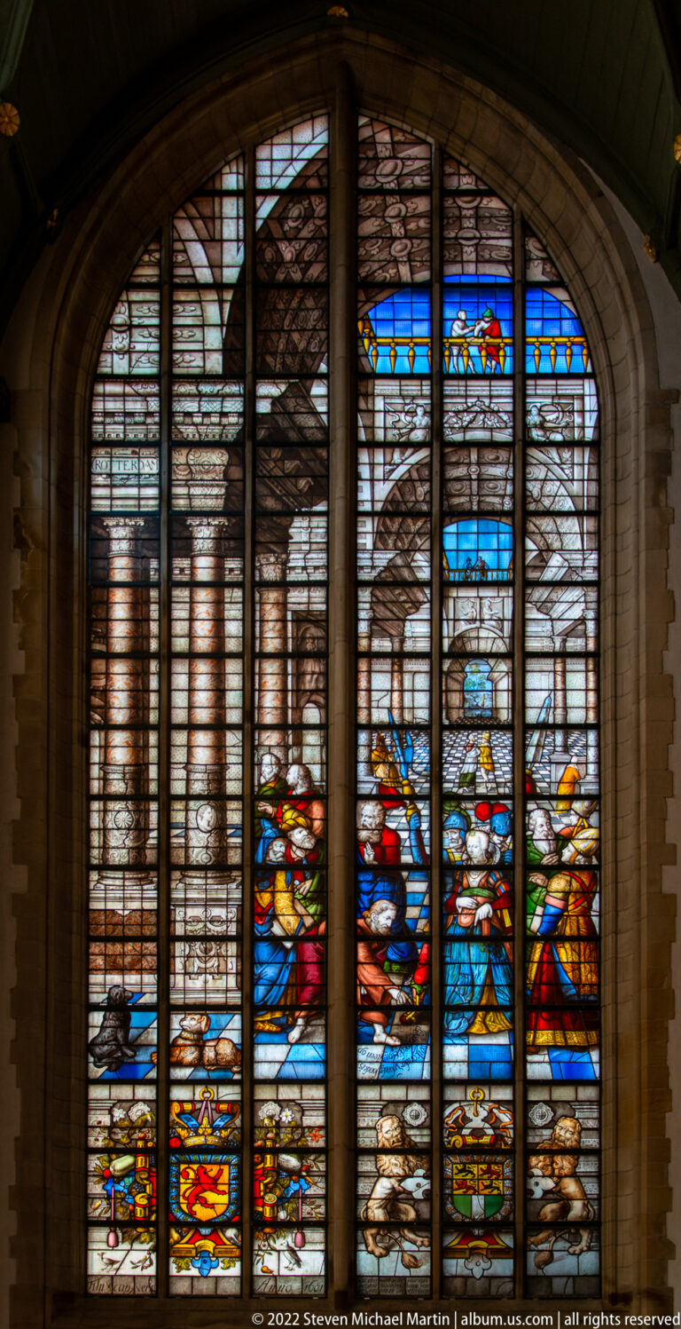 Arched stained-glass window with pale architectural scenes, colorful figures, and heraldic details below.