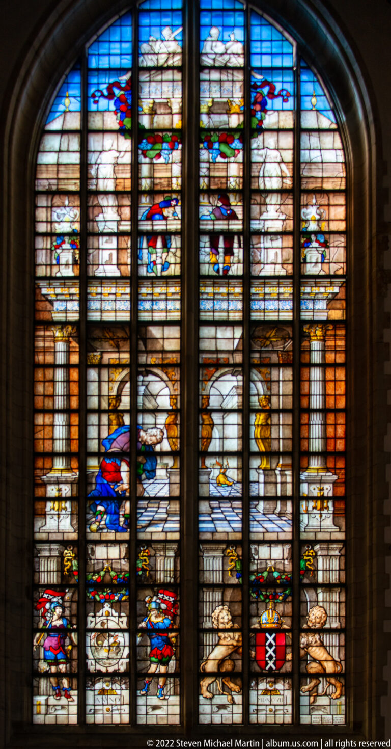 Tall stained-glass window with classical architecture, small figures, and colorful heraldic details.