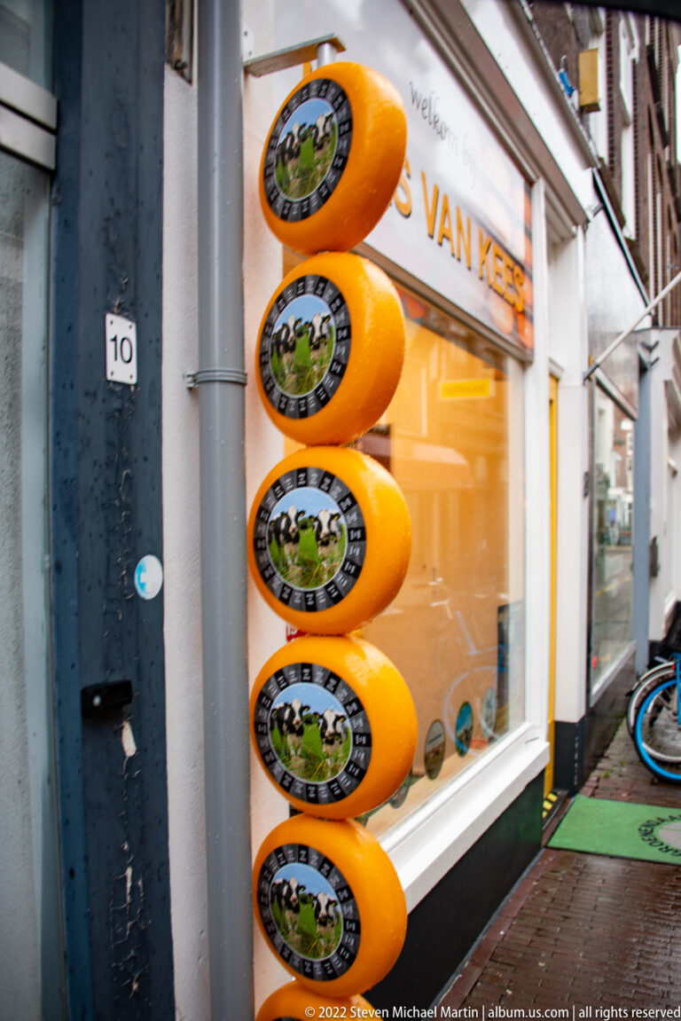 Five orange cheese-wheel signs stacked vertically beside the entrance and window of a Gouda cheese shop.