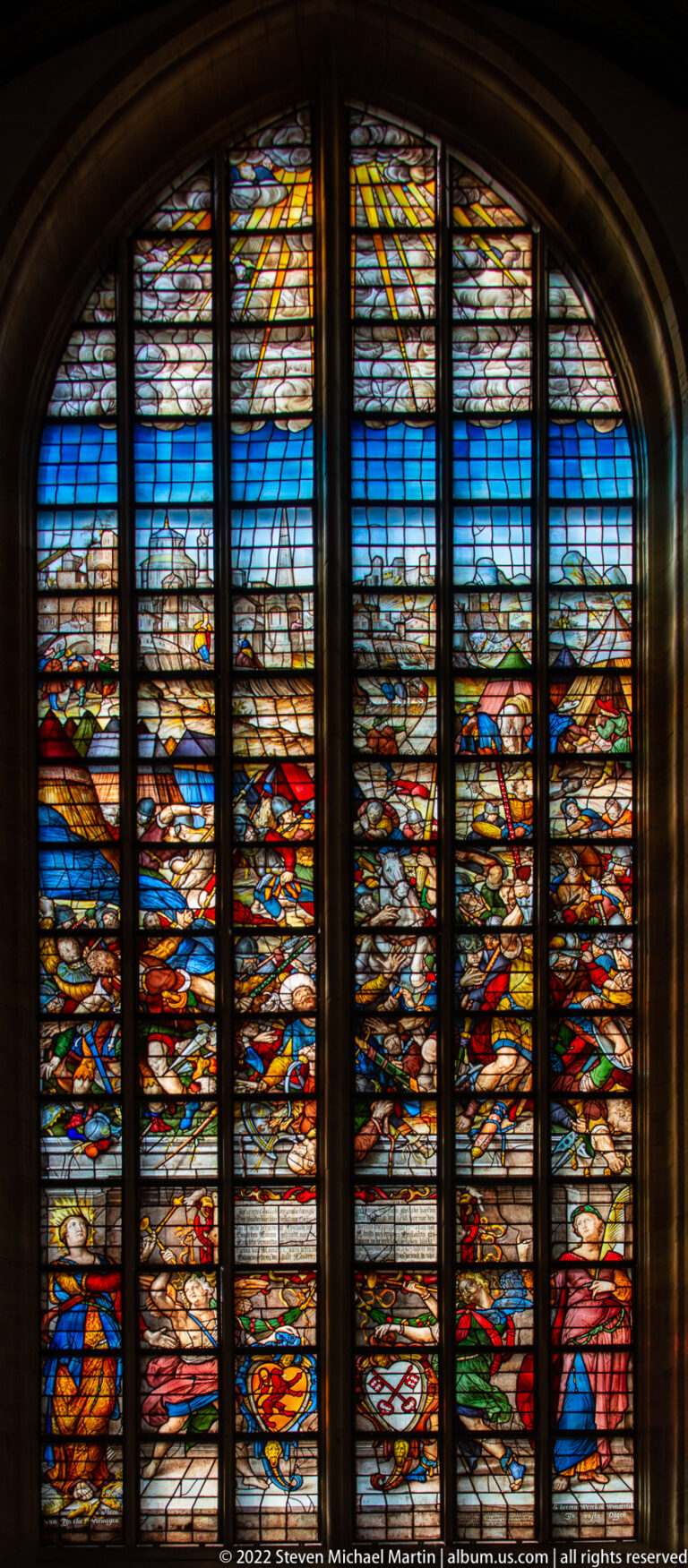 Wide stained-glass scene crowded with mounted and standing figures, banners, and weapons beneath blue sky.