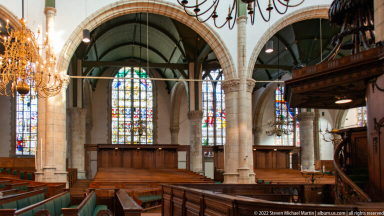 Side interior of Sint-Janskerk with wooden benches, stone columns, chandeliers, and tall stained-glass windows.