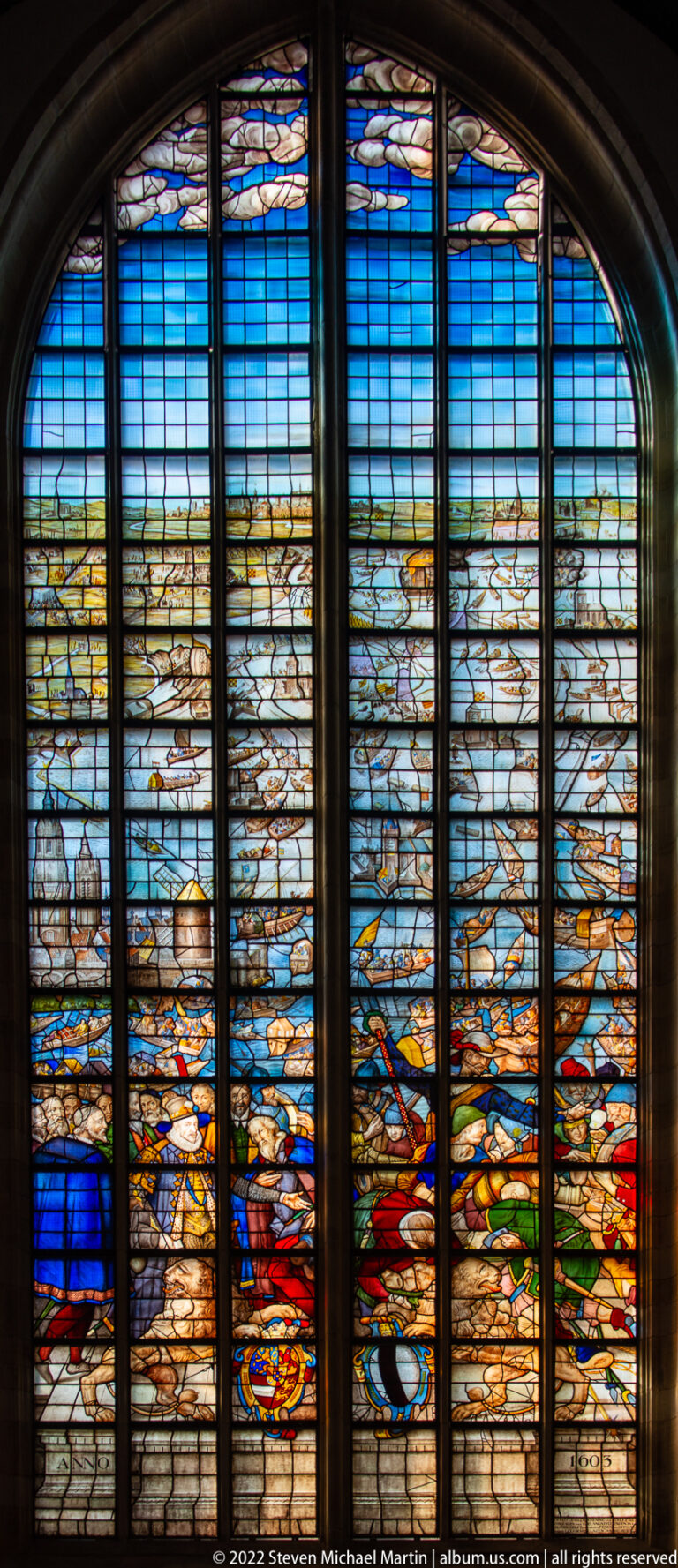 Wide stained-glass landscape with blue water or sky, distant buildings, boats, and figures across several panels.