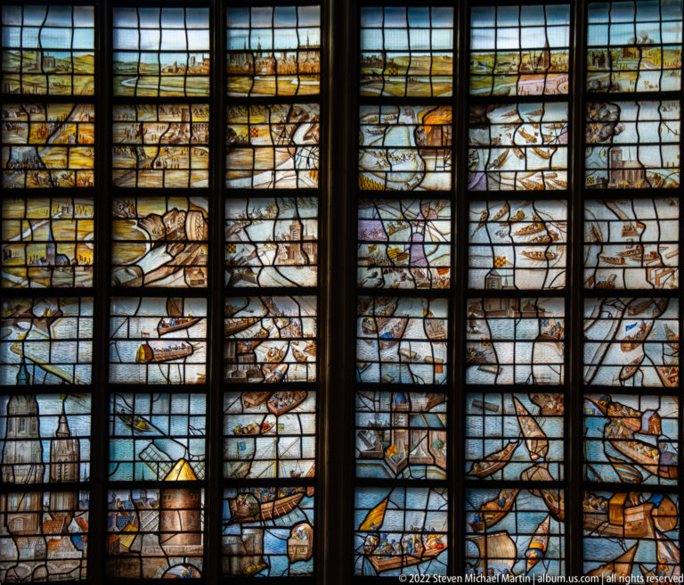 Stained-glass maritime scene with sailing ships, small boats, shoreline buildings, and figures.