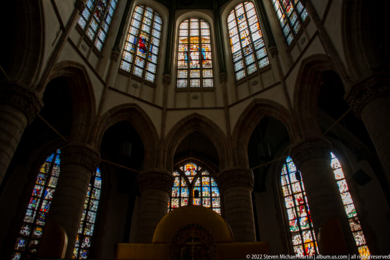 Curved church interior with stone columns, pointed arches, and tall stained-glass windows above.