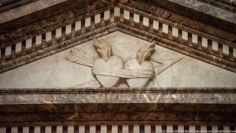 Marble relief of a flaming heart with wing-like forms set within a triangular architectural field.