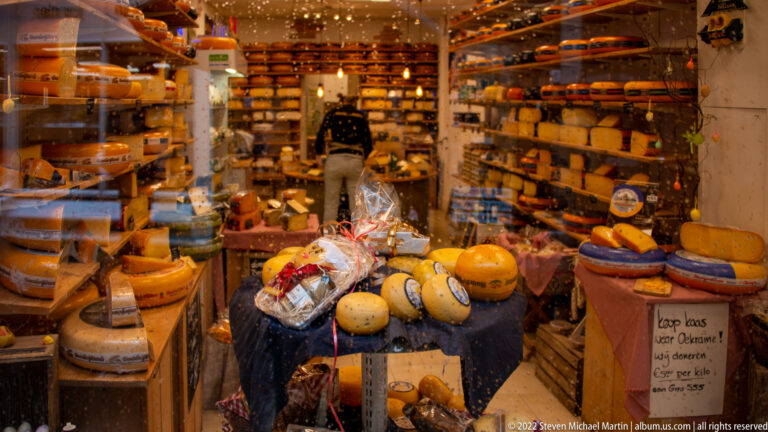 Warmly lit cheese shop packed with wheels, wedges, packaged cheeses, display tables, shelves, and a staff member.