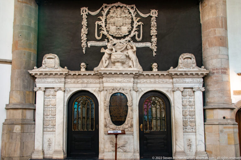 Large white marble wall monument with two dark doors, inscription tablet, skulls, figures, and heraldic crest.