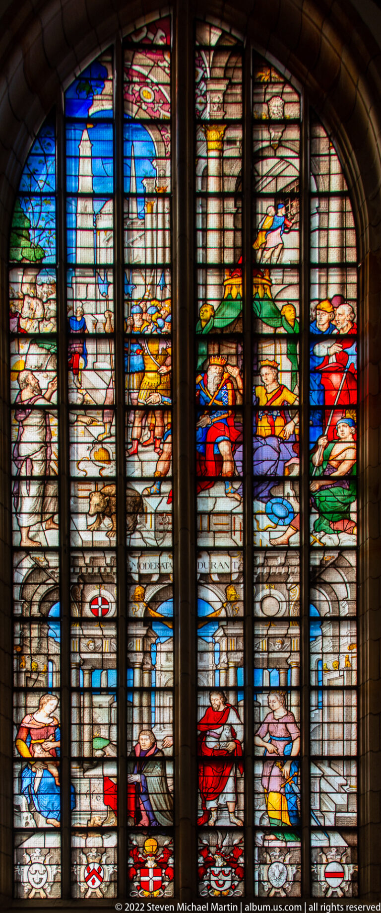 Stained-glass window with colorful figures arranged against an architectural city and interior setting.