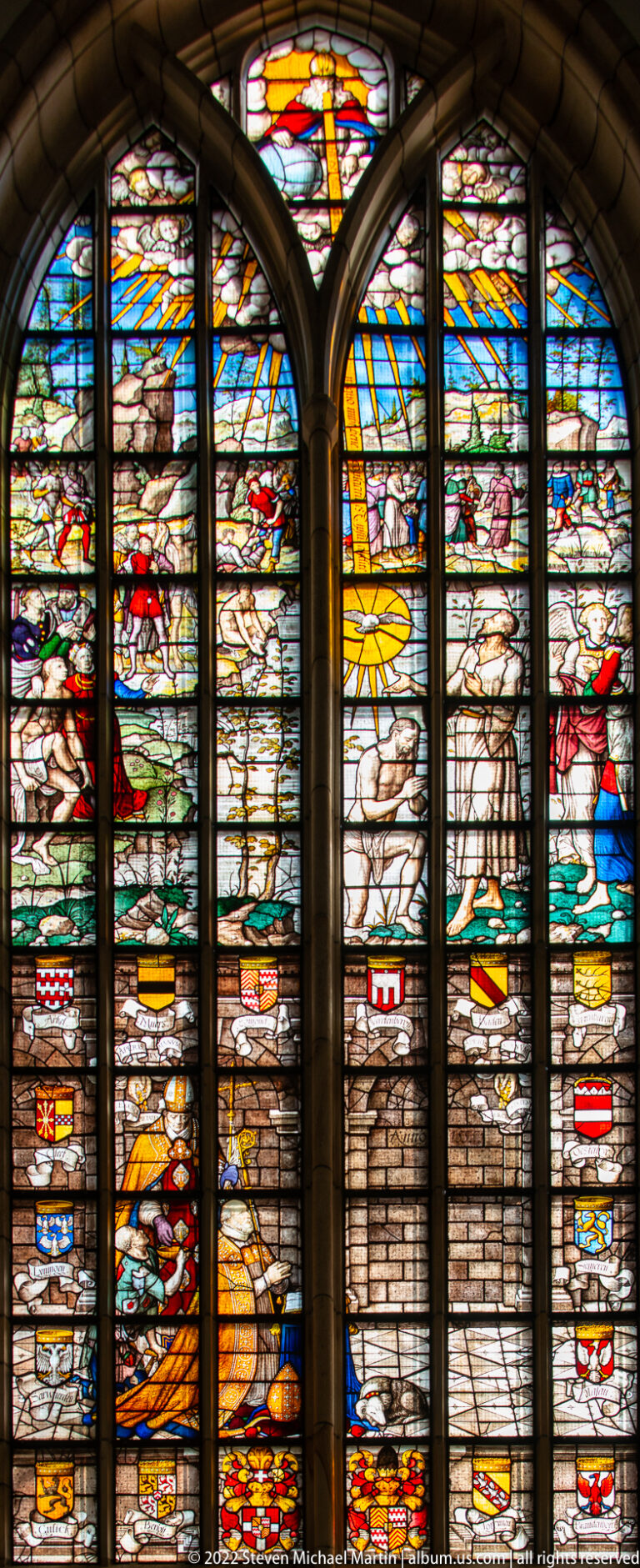 Paired-arched stained-glass window with radiant yellow upper scene, figures, angels, and heraldic details.