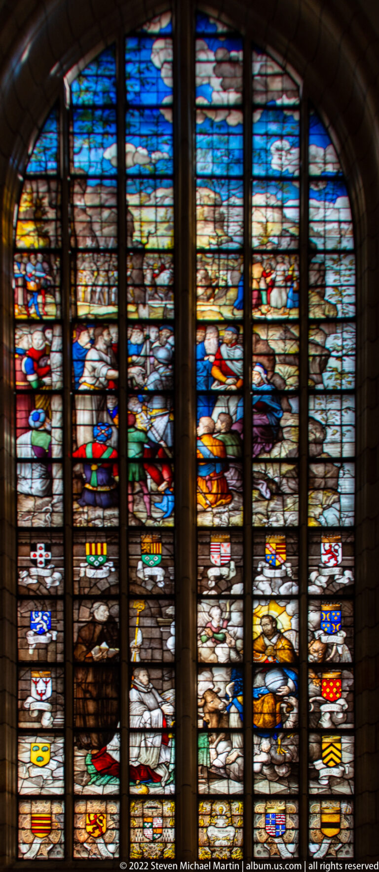 Large stained-glass window with narrative scenes, colorful figures, and many heraldic shields.
