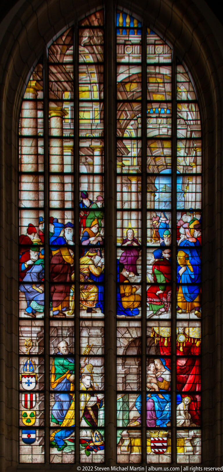 Arched stained-glass window with richly dressed figures in groups and heraldic shields along the lower panels.