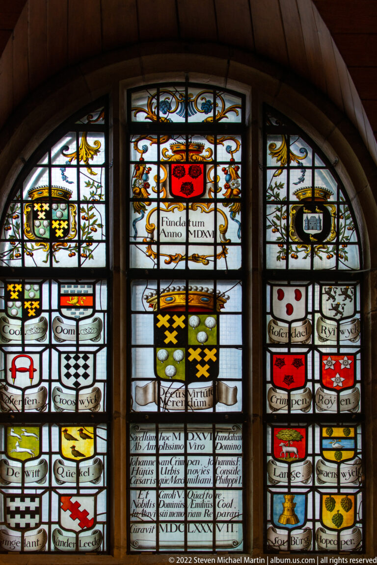 Arched stained-glass window filled with coats of arms, inscribed scrolls, and a larger central heraldic shield.