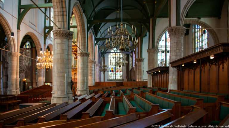 Wide church interior with wooden benches, stone arcades, green timber roof, brass chandeliers, and stained glass.