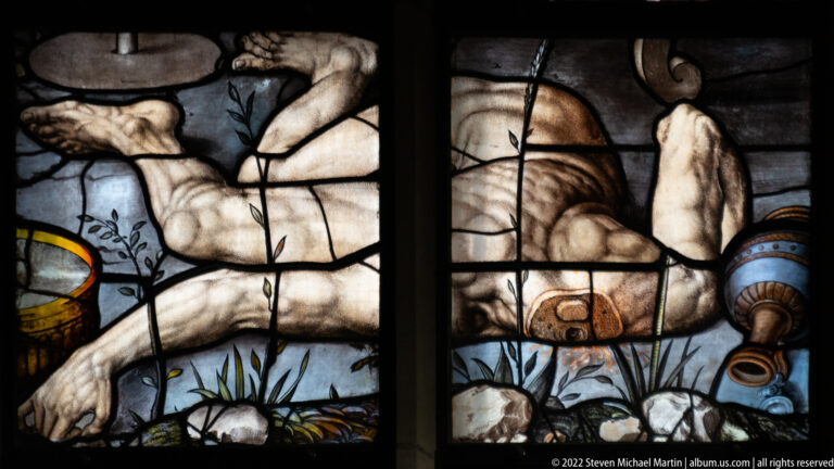 Close stained-glass detail of a fallen nude figure beside a tipped vessel and small plants.